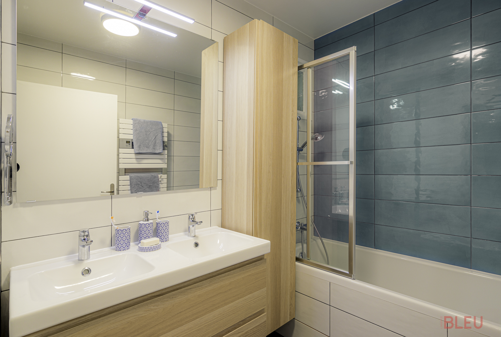 La rénovation de la salle de bain implique le remplacement de la grande baignoire par une baignoire de taille standard, permettant ainsi de libérer de l'espace. Un double plan vasque est installé avec un pare-baignoire pour éviter les éclaboussures.