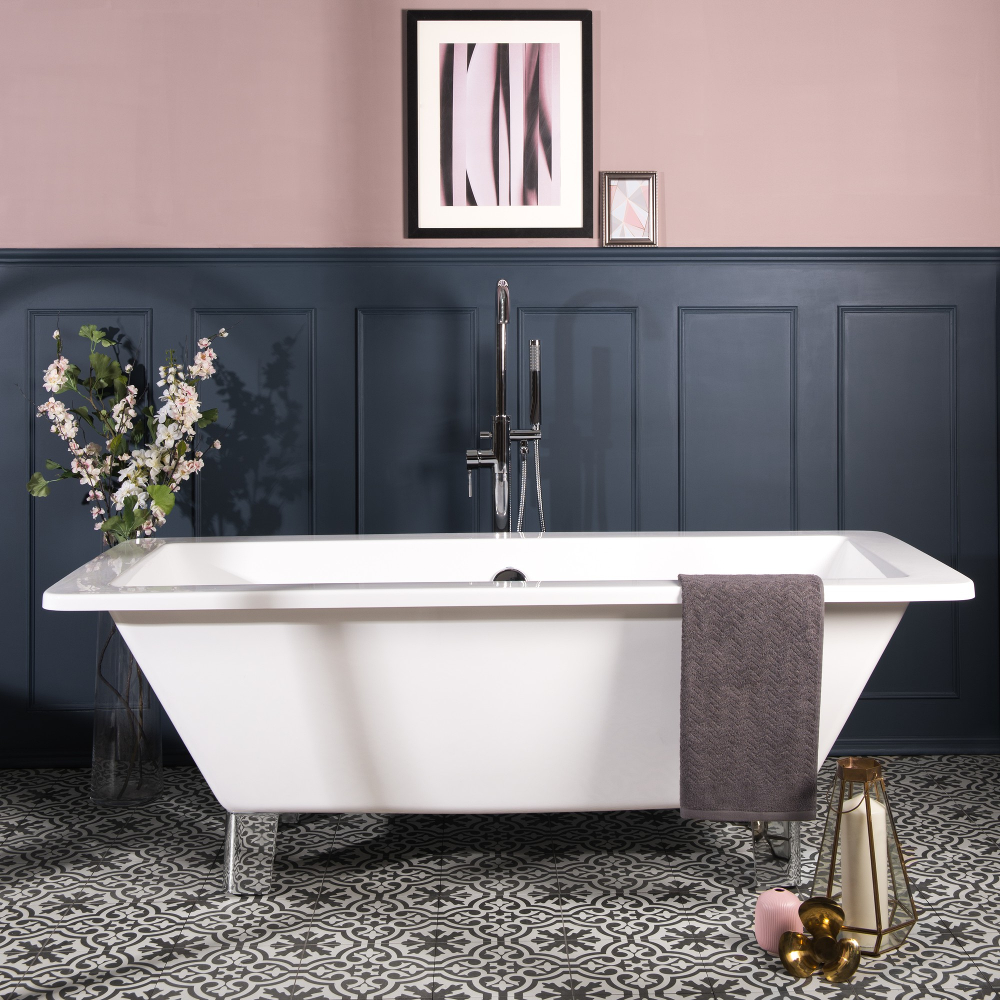 L'Agence BLEU, salle de bain élégante et colorée. La pièce est dominée par une baignoire îlot blanche positionnée au centre sur un sol en carreaux de céramique aux motifs géométriques en teintes de rose pastel et bleu foncé. Les murs sont peints d'un bleu profond qui crée un contraste audacieux avec le rose du sol. Une petite table d'appoint blanche est placée à côté de la baignoire, idéale pour poser des objets de bain. La pièce est éclairée par une lumière douce et indirecte, renforçant l'atmosphère relaxante de la salle de bain. Le design général est à la fois moderne et féminin, avec une touche vintage apportée par le sol à motifs.