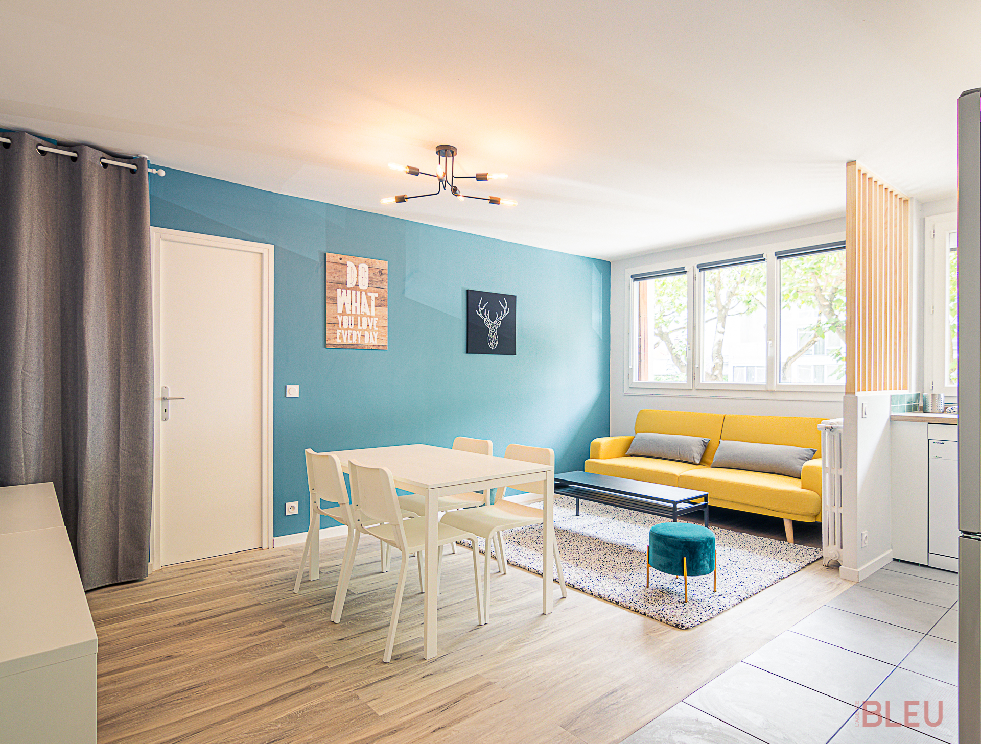 Vue d'un espace de vie moderne et lumineux, optimisé par l'Agence BLEU, cabinet d'architecture Paris et architecte d'intérieur Paris. Comprend un coin repas avec une table blanche et un coin salon avec un canapé jaune vif contre un mur bleu turquoise